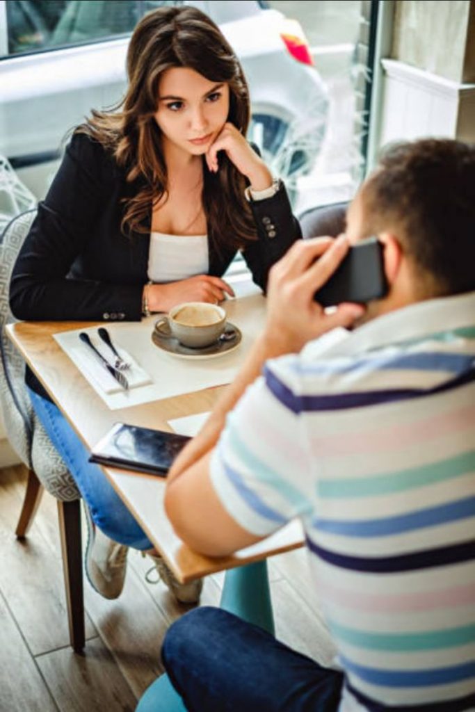 Casal na cafeteria, namorada irritada porque o namorado fala com outra pessoa no celular.