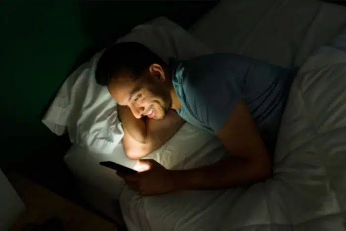 Homem sorridente com o celular na mão deitado na cama, ouvindo o que ele queria ouvir.