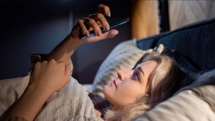 Moça com o celular na mão na cama mandando mensagens de texto picantes