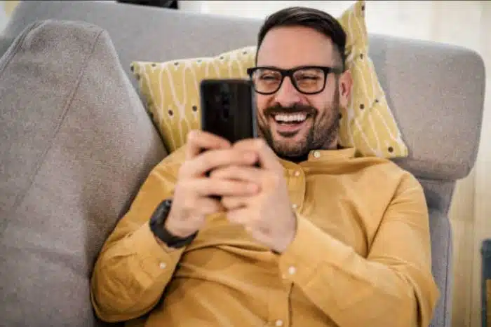 Homem sorridente deitado no sofá te mandando mensagem pelo celular